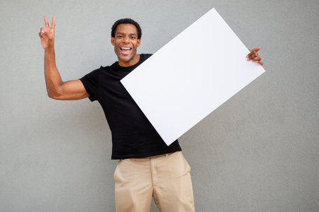 A man in a black t-shirt holds a blank white poster board and flashes a peace sign, standing against a grey wallの写真素材