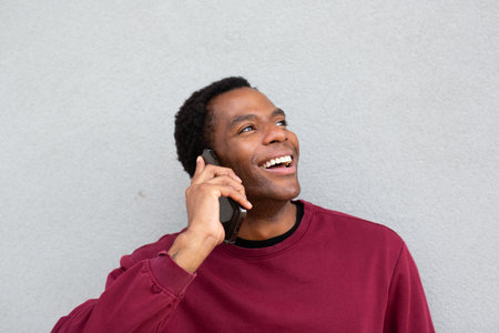 Man in a red sweatshirt standing against a gray wall, looking up and smiling while talking on his smartphoneの写真素材
