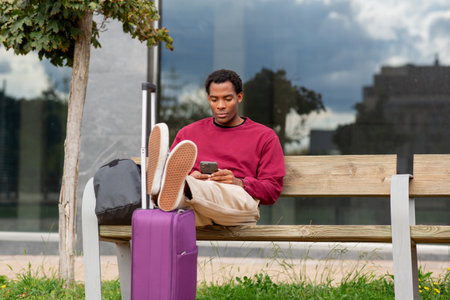 A man in a burgundy sweatshirt and beige pants sitting on a bench with his feet resting on a purple suitcase, using a smartphoneの写真素材
