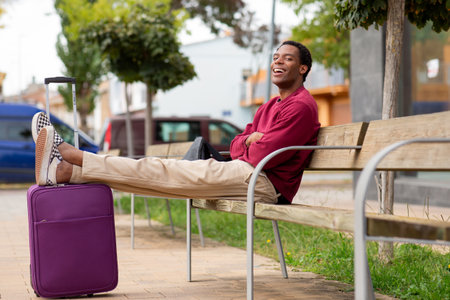 smiling man sitting on a wooden bench with legs resting on a purple suitcase, wearing a red sweater and beige pantsの写真素材