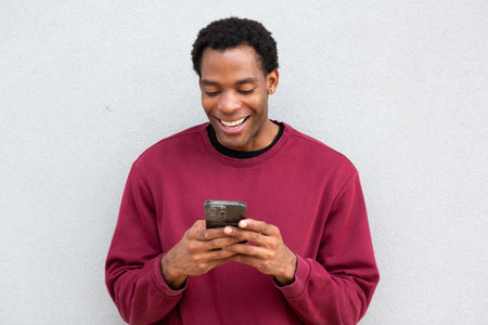 Man in a red sweatshirt standing against a gray wall, smiling and looking at his smartphone. He is using both hands to hold the phoneの写真素材