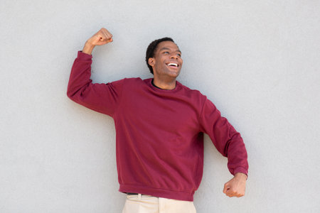 A man wearing a red sweater flexes his arm while smiling, standing in front of a grey wall