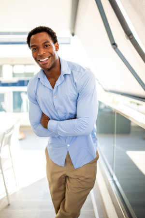 A smiling man dressed in a blue shirt and beige pants stands with his arms crossed in a modern, sunlit spaceの写真素材