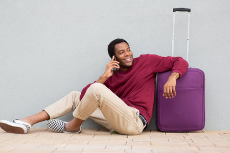 A man in a burgundy sweatshirt and beige pants sitting on the ground next to a purple suitcase, smiling while talking on the phoneの写真素材