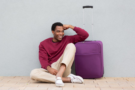 Man sitting on a tiled sidewalk next to a purple suitcase, smiling while resting against a wall. He is wearing a burgundy sweater, beige pants, and checkered shoes, with a plain grey backgroundの写真素材
