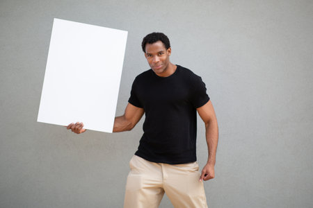 Man wearing a black t-shirt and beige pants holding a blank white sign in one hand with a confident expressionの写真素材