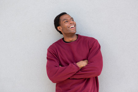 A man in a red sweater stands with his arms crossed and looks upward, smiling, in front of a grey wallの写真素材