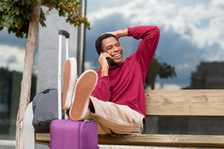 Man sitting on a wooden bench outdoors, resting his legs on a suitcase and talking on a smartphone. He smiles while holding his head with one hand, next to a black backpack and purple suitcaseの写真素材