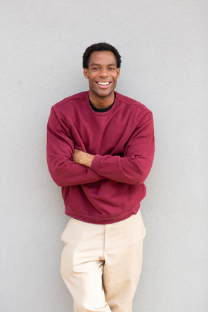 A man smiling with his arms crossed, wearing a red sweater and beige pants, standing against a gray wallの写真素材