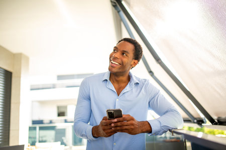 Man wearing a light blue shirt, holding a smartphone and smiling while standing on a balcony, with a modern building and bright lighting in the backgroundの写真素材