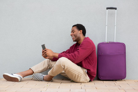 A man wearing a burgundy sweatshirt and beige pants sitting on the ground next to a purple suitcase, smiling while using a smartphoneの写真素材