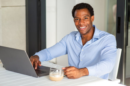 A man in a blue shirt smiling while working on a laptop at a table with a glass cup of coffeeの写真素材