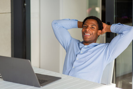 A man in a blue shirt smiling and leaning back with his hands behind his head during a break from working on his laptop at a tableの写真素材