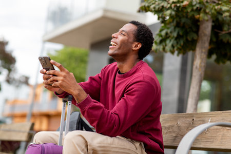 Happy man holding a smartphone and laughing while sitting on a wooden bench with a purple suitcase beside himの写真素材