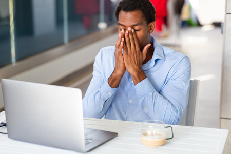 Frustrated man in a blue shirt sitting at a table with a laptop and covering his face with his handsの写真素材