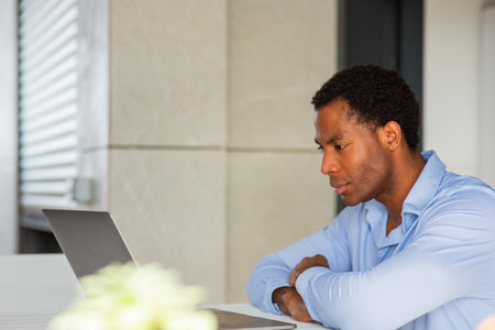 A man in a blue shirt deeply focused on his laptop at a tableの写真素材