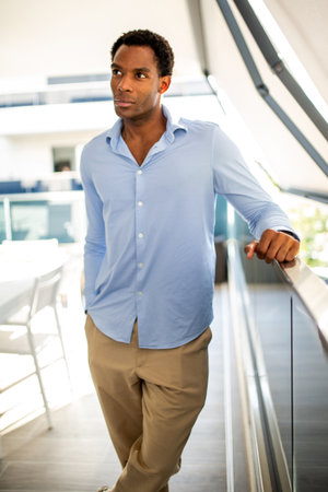Man dressed in a blue shirt and beige pants, standing by a railing in a bright, modern spaceの写真素材