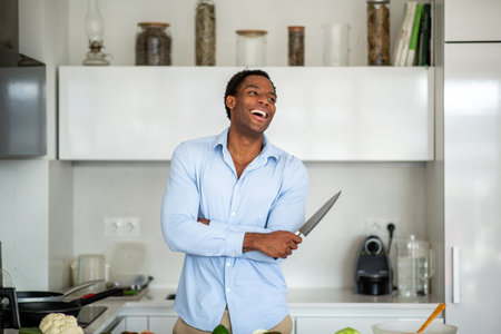 Laughing man dressed in a blue shirt and beige pants standing in a modern kitchen while holding a knifeの写真素材