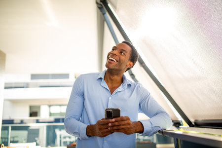 Man in a light blue shirt, smiling and holding a smartphone, standing on a balcony in an outdoor setting with modern architecture in the backgroundの写真素材