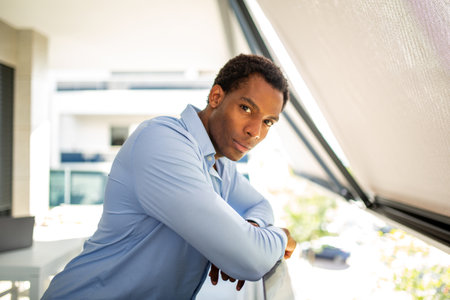 A man in a blue shirt standing on a balcony, leaning on the railing while looking out thoughtfullyの写真素材