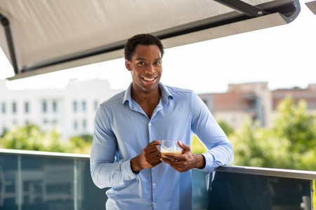 Man in a light blue shirt smiling and holding a cup while leaning on a balconyの写真素材