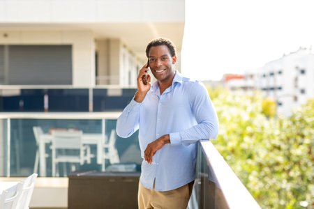 Smiling man in blue shirt talking on his smartphone while leaning on a balcony railing with blurred buildings and greenery in the backgroundの写真素材