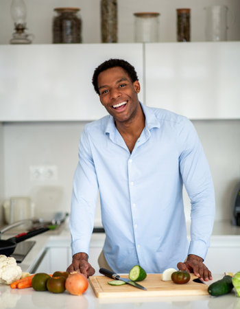 Smiling man wearing a blue shirt leaning over a cutting board while preparing vegetables in a modern...