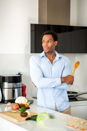 Man wearing a blue shirt and beige pants holding a wooden spoon while standing in a modern kitchenの写真素材