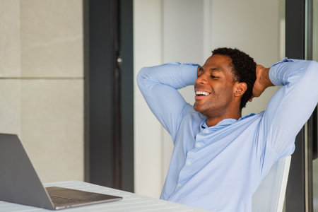 A man in a blue shirt sitting back and smiling with his hands behind his head after working on a laptop at a tableの写真素材