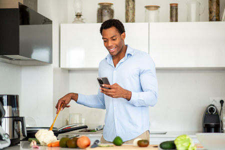 Man multitasking in the kitchen, using a wooden spatula to cook while checking his smartphoneの写真素材