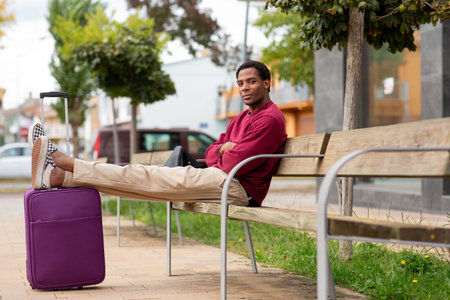 Man sitting on a wooden bench with his legs resting on a purple suitcase, wearing a red sweater, beige pants and checkered shoesの写真素材