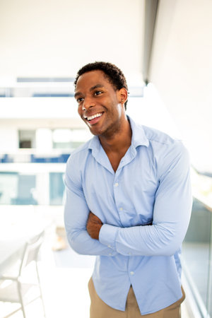 A laughing man in a blue shirt and beige pants stands with his arms crossed in a bright, modern environmentの写真素材