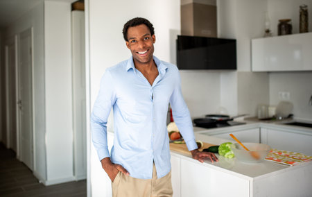 Smiling man dressed in a blue shirt and beige pants standing next to a kitchen counter with vegetables and kitchen tools laid outの写真素材