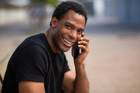 A man in a black t-shirt smiling and holding a smartphone to his ear, engaged in a phone conversation while sitting on a bench in an outdoor settingの写真素材