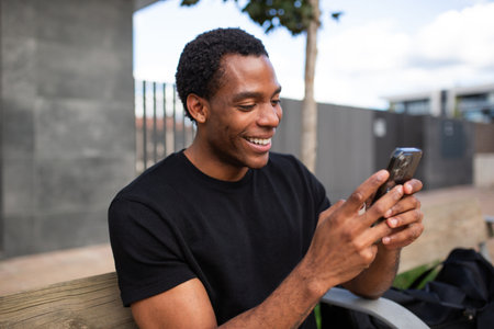 Man sitting on a wooden bench outdoors, smiling as he looks at his smartphoneの写真素材