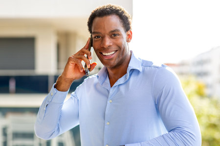 Smiling man in blue shirt holding a smartphone to his ear while standing outdoors on a rooftop with blurred buildings and greenery in the backgroundの写真素材