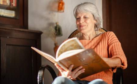 A senior woman with grey hair is seen smiling as she enjoys reading a book in a warmly lit roomの写真素材