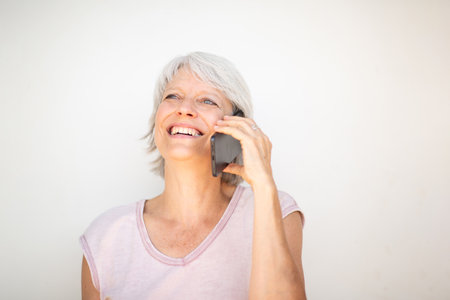 Portrait of a cheerful mature woman with short gray hair, holding a smartphone to her ear while smiling and looking slightly upwardの写真素材