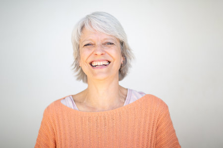 Close-up portrait of a mature woman with short gray hair, wearing an orange sweater, smiling warmly in front of a white backgroundの写真素材