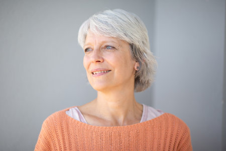 Portrait of a mature woman with short gray hair, wearing an orange sweater and looking thoughtfully to the side, standing against a light gray backgroundの写真素材