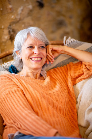 A cheerful senior woman with grey hair and a bright smile, relaxing on a couchの写真素材