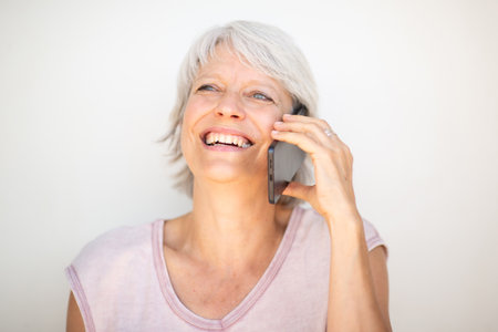 Close-up portrait of a joyful mature woman with short gray hair, holding a smartphone to her ear while smiling widelyの写真素材