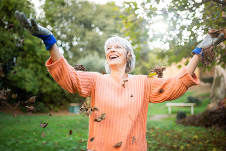 Happy elderly woman dressed in an orange sweater and gloves, throwing autumn leaves and smiling in the gardenの写真素材
