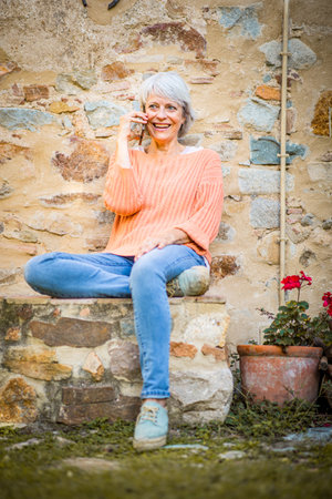 A senior woman with grey hair is seated outdoors, talking on a smartphone. She is wearing an orange sweater and jeans, sitting casually against a rustic stone wallの写真素材
