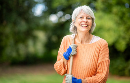 Senior woman with short gray hair, wearing an orange sweater and blue gardening gloves, smiling while holding a rake in an outdoor garden settingの写真素材