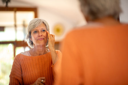 Senior woman in an orange sweater applying skincare to her face while looking in the mirrorの写真素材