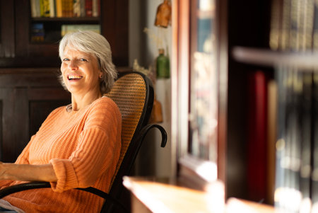 Mature woman with gray hair, dressed in an orange sweater, smiling and relaxing in a cozy chair in her home libraryの写真素材