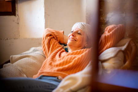 Smiling elderly woman in an orange sweater reclining with hands behind her head on a comfortable, cushioned chairの写真素材