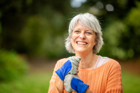 Senior woman with short gray hair, wearing an orange sweater and blue gardening gloves, smiling while standing in a gardenの写真素材