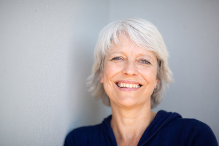 Close-up portrait of a mature woman with short gray hair, wearing a navy blue jacket and leaning against a gray wall with a joyful smileの写真素材